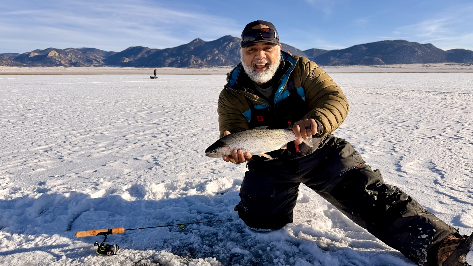 Anthony Jimenez kneeling on a frozen Colorado reservoir, holding up a chrome rainbow trout with snow-covered peaks behind him