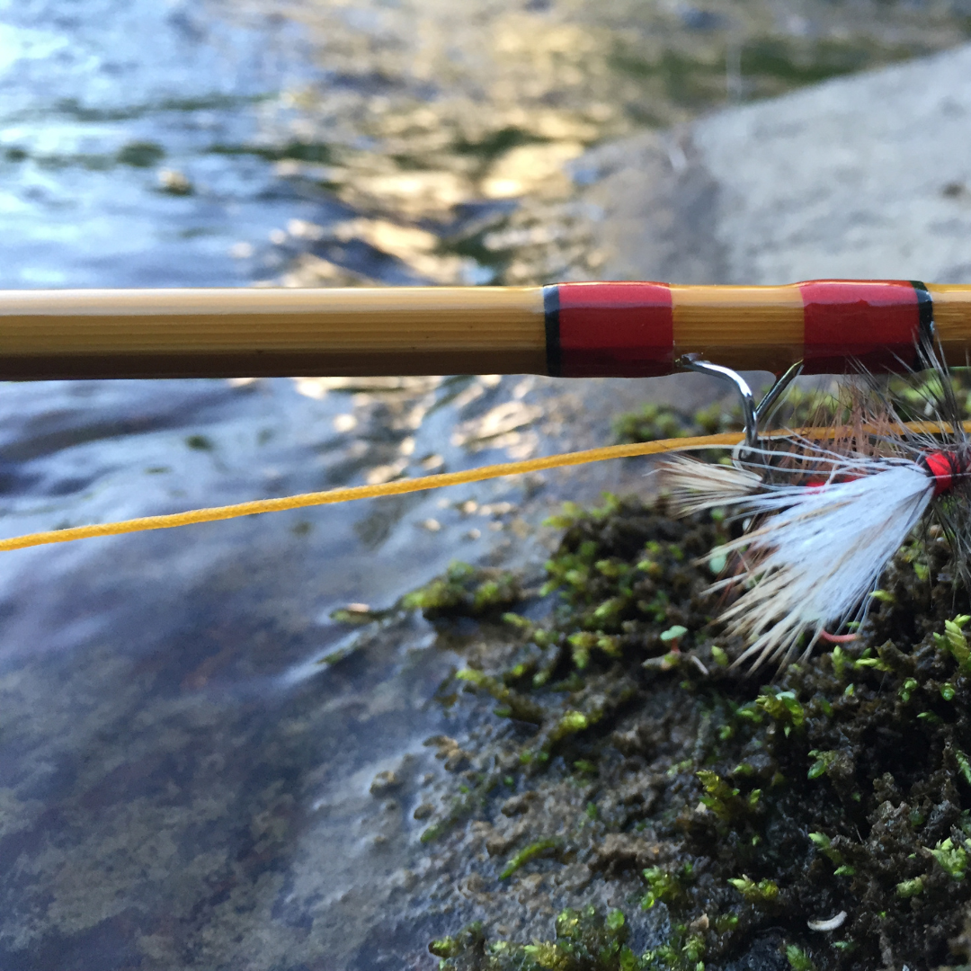 A hand-planed bamboo blank with red and black thread wraps, a white-winged dry fly hooked on the snake guide and yellow fly line running off over a mossy rock and shallow moving water (photo: Headwaters Bamboo)