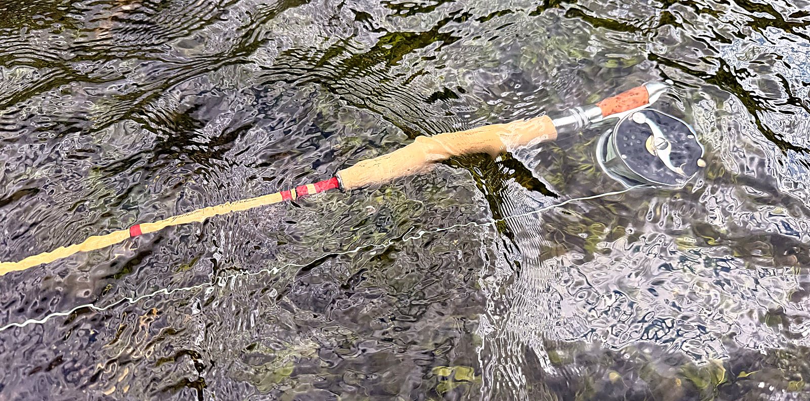 The Headwaters Gallatin lying in clear shallow water over cobble — blonde cane with red thread wraps, cork grip, knurled reel seat and the Clark Fork click-and-pawl, fly line trailing through the current
