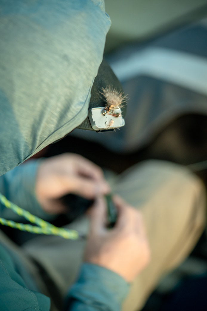 A silver Stash Tray clipped to a green cap brim holding a single dry fly, with an angler's hands tying on tippet in soft focus below (photo: Lid Rig)